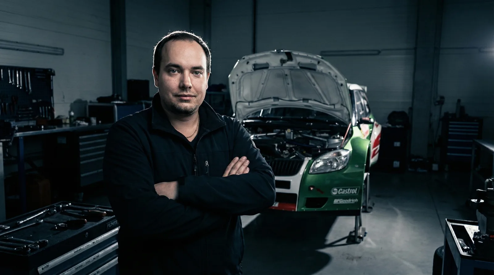 Jiří Caha se založenýma rukama v dílně, za ním zelenobílá Škoda Fabia S2000 s otevřenou kapotou